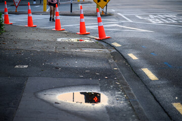 Orange traffic cones lining up the street. Red traffic arrow sign reflected in the puddle of...