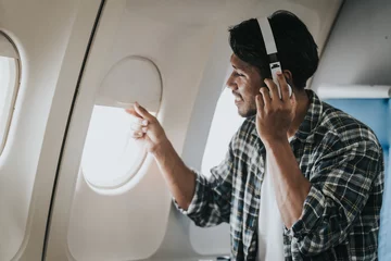 Fotobehang Asian man on an airplane looking out the window with headphones on, possibly enjoying music or an audiobook during his flight. © Ratirath
