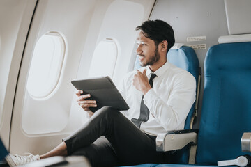 Asian businessman in an airplane, intently engaged with a tablet, potentially working or reading while traveling.