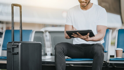 Asian man sitting in an airport lounge, focused on a tablet, with a suitcase next to him, portraying him as a traveler or a professional on the go.