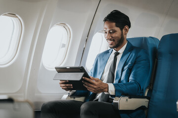 Fototapeta premium Asian businessman working on his laptop while seated in an airplane, smiling, possibly engaged in work or leisure during his flight.