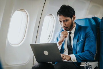 Asian businessman working on his laptop while seated in an airplane, smiling, possibly engaged in work or leisure during his flight.
