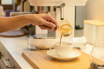 Woman hand pouring fresh hot espresso coffee into a white cup in the kitchen at home in the morning
