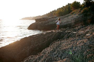 Young traveler woman in white shirt and shorts walking on the rock near the sea with amazing sunset view