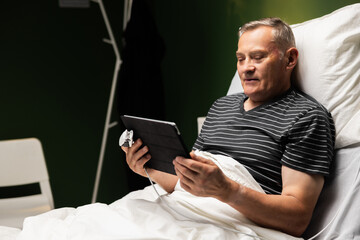A senior patient in the hospital relaxes, using his tablet in bed. He browses various websites,...