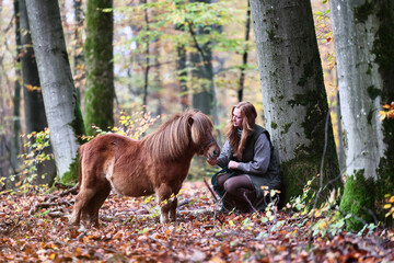 Young woman with long red hair in a winter coat and knitted dress with her small horse (Shetland...
