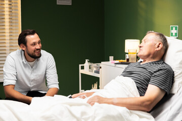 A happy old man enjoys visiting his son during his recovery in a hospital bed.