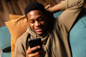 Happy delighted attractive African music lover, streaming his favorite tunes on his phone while...