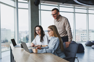 Three employees in a modern office, a man and a woman work at a table, colleagues discuss and...
