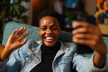 Portrait of delighted afo american dark skinned woman blogger vlogger taking selfie photo on camera sitting at home at weekends in cozy living room making stream online live. - obrazy, fototapety, plakaty