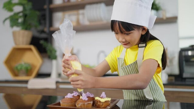 Little toddler asian girl child in apron and chef hat whipped cream decorating preparing homemade cupcakes in home kitchen. A Little girl preparing and decorating homemade cake. Children cooking