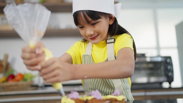 Little toddler asian girl child in apron and chef hat whipped cream decorating preparing homemade cupcakes in home kitchen. A Little girl preparing and decorating homemade cake. Children cooking