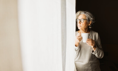 Thoughtful senior woman standing by a window and drinking coffee on a sunny day at home. - obrazy, fototapety, plakaty