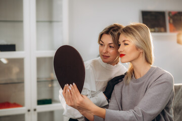 Bottom view of mirror reflection of smiling caucasian girl looking at her beautiful lips while female beautician looking at her after permanent makeup lips procedure in beauty salon. - obrazy, fototapety, plakaty