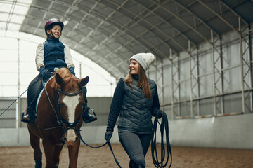 Young woman, teacher, instructor teaching little girl horseback riding. Child sitting in paddle, practicing. Education. Concept of sport, childhood, school, course, active lifestyle and hobby - obrazy, fototapety, plakaty