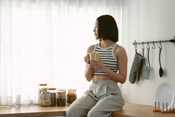 Portrait of beautiful Asian woman holding hot coffee and looking through window while sitting on top counter kitchen. Korean or Japanese girl relax on cozy at home. Lifestyle and Relax time concept.