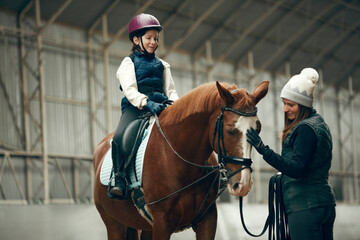 Female instructor teaching little girl, child horseback rising. Kid practicing trail riding,...