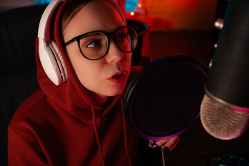 Portrait of a womam wearing headphones in a red hoodie and glasses recording a podcast live into a...