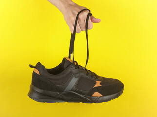 Hand holding a black sneaker by the laces on a yellow background