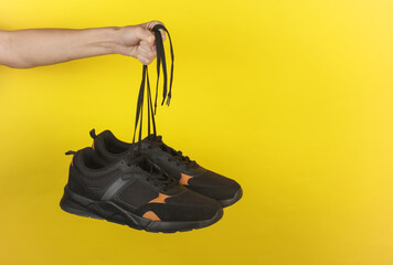 Hand holding a black sneakers by the laces on a yellow background - Powered by Adobe