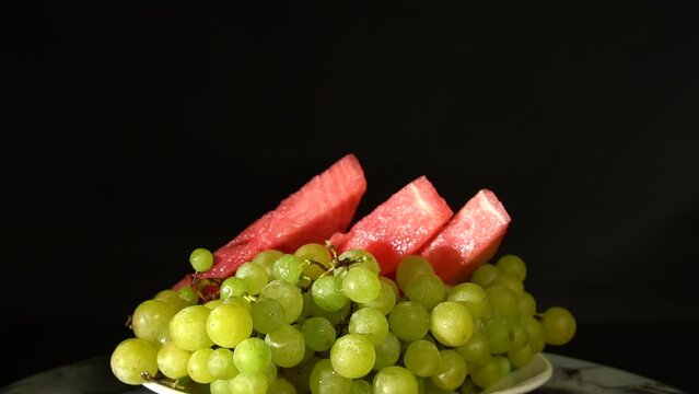 Ripe green grapes and slices of sweet, juicy watermelon on a black background. Ideal for content about summer food and healthy lifestyle. 4k footage.