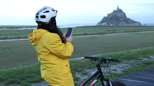 woman with raincoat and bicycle taking a photo at the Mont Saint Michel monument, France