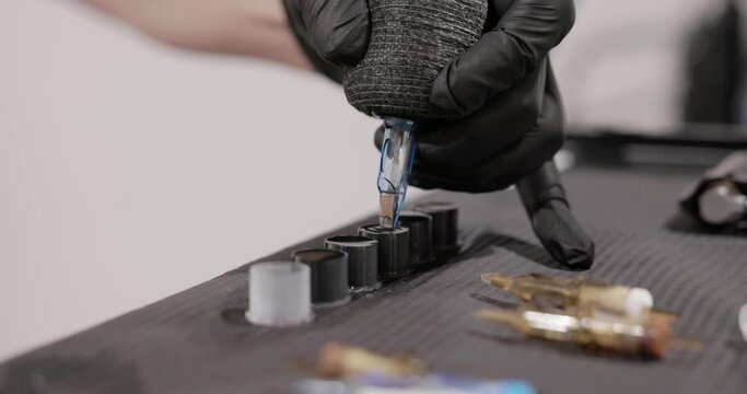 Tattoo Artist Dips The Tip Of The Needle Cartridge Into A Filled Ink Cap On The Table. - close up shot