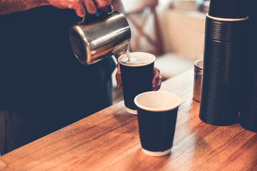 Crop barista pouring hot milk from iron mug into coffee cups