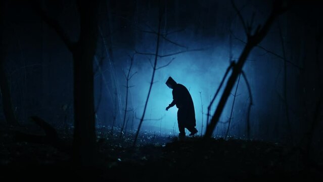 Mysterious dark figure in the night foggy forest. Silhouette of an unrecognizable man in a tunic with a hood.