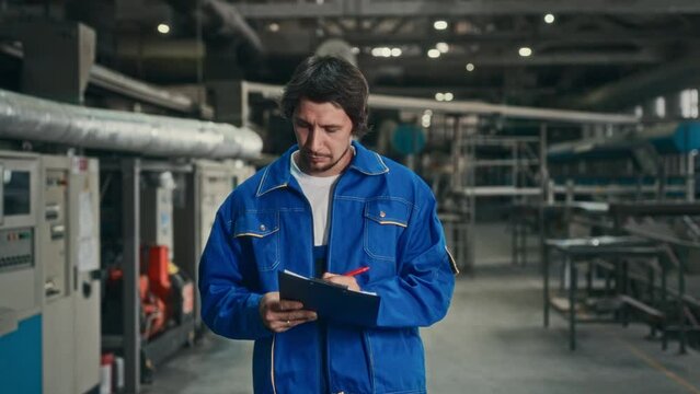 A male engineer in a blue uniform walks through the factory workshop and checks the operating parameters of an automatic production line. Work on setting up automatic equipment.