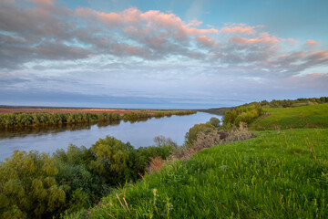A river against a backdrop of green fields, a breathtaking view from the hill. Colorful sky with clouds during sunset. Landscape in spring. Nature during a bright sunset.
