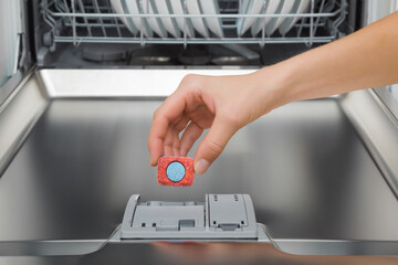Young adult woman hand fingers holding and adding blue red dishwasher tablet in detergent dispenser for dishes washing. Closeup. Front view. - obrazy, fototapety, plakaty