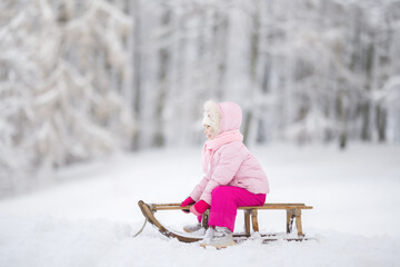 Happy little girl in pink warm clothes sitting on sledge and sledding down on snow from hill. Child...