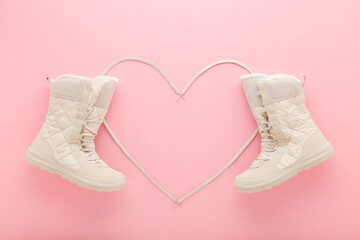 Heart shape created from white shoelaces between new beige warm winter boots on light pink table background. Pastel color. Female footwear. Closeup. Top down view.