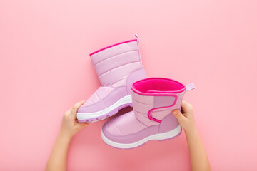 Little girl hands holding new warm waterproof winter boots on light pink table background. Pastel color. Children footwear. Closeup. Point of view shot. Top down view. - obrazy, fototapety, plakaty