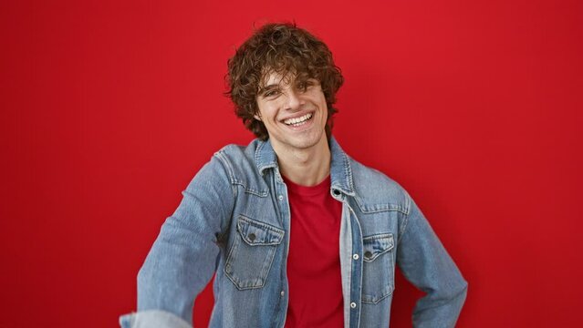 A happy young adult man with curly hair, wearing a red shirt and denim jacket, gives a thumbs up against a vibrant red background.