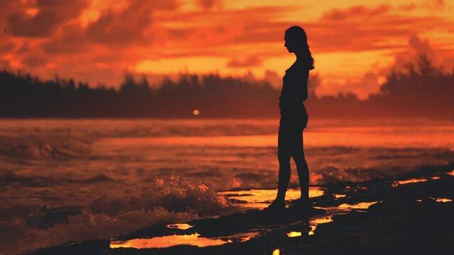 Dramatic bloody red sunset over ocean shore beach. Slim woman silhouette standing on stony coastline watching splashing storm waves tropical landscape. Female tourist on summer holiday vacation.