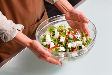 Female hands holding a glass bowl of vegetable salad with feta over a glass table.