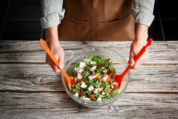 Female hands mixing a vegetable salad in a glass bowl against a wooden background - obrazy, fototapety, plakaty