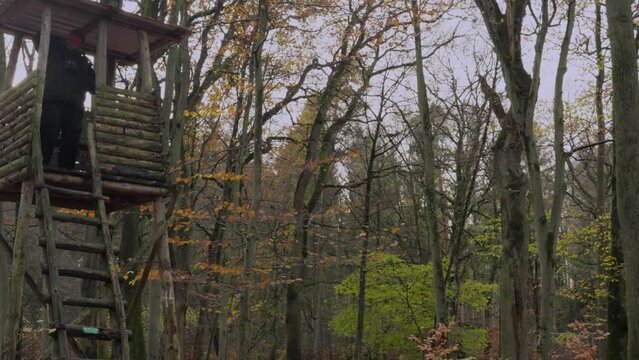 A hunter in a thick winter suit sits on a high stand, then gets up and climbs down the ladder and runs out of the picture, he carries a hunting rifle with a silencer over his shoulder.