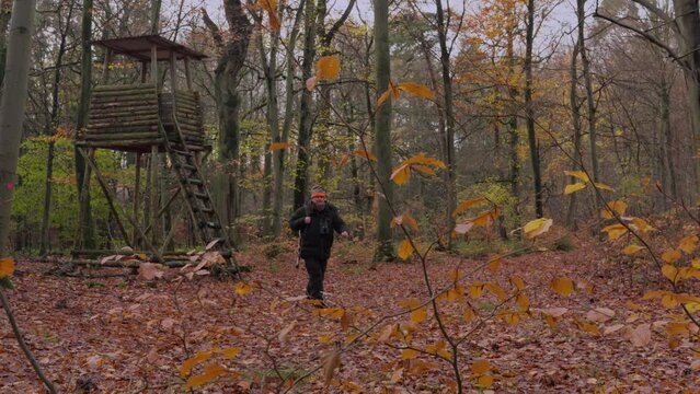 Long shot of a hunter walking away from a high stand in a thick winter suit, carrying a hunting rifle with a silencer over his shoulder. The camera looks through the bushes.