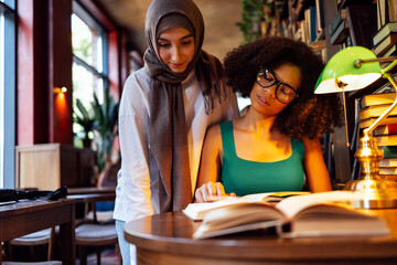 Two teenage girls of different nationalities are in the library and read books. African american girl with glasses shows muslim girl in hijab to page.