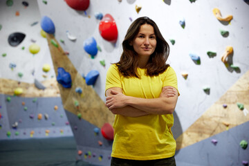 Fototapeta premium Portrait of young smiling woman in yellow t-shirt, bouldering instructor standing with climbing wall behind. Concept of bouldering, sport climbing, hobby, active lifestyle, school, training course