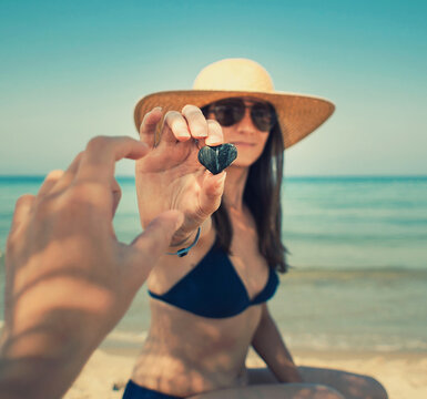 Love concept, a young woman is holding a heart-shaped pebble and holding it in front of her boyfriend. Two lovers at the beach