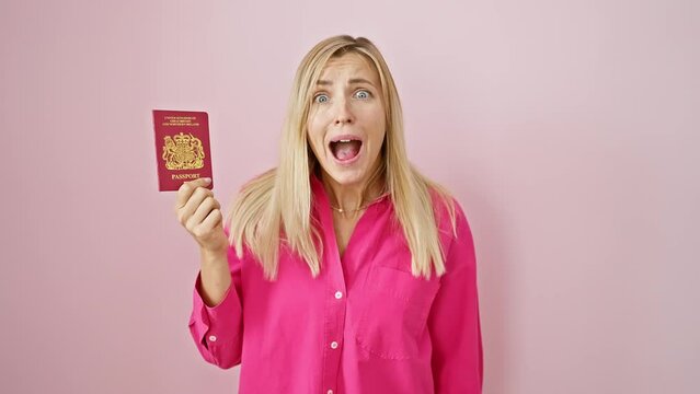 Blonde young woman, visibly freaked, staring with open-mouthed disbelief while clutching her north ireland passport over isolated pink background.