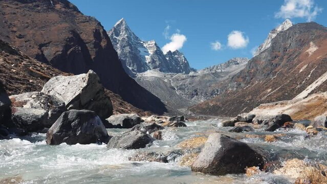 Clear stream running over stone boulders in highlands of Himalayas. River flowing along stone bottom against background rocky snowy mountains. Water of mountain river splashes on sunny summer day