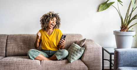 Excited happy young black woman holding smart phone device sitting on sofa at home - Happy satisfied female looking at mobile smartphone screen gesturing yes with clenched fist - Technology concept - obrazy, fototapety, plakaty