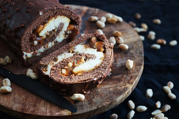 Chocolate roll with cream and caramel next to peanuts on a wooden tray on a dark background