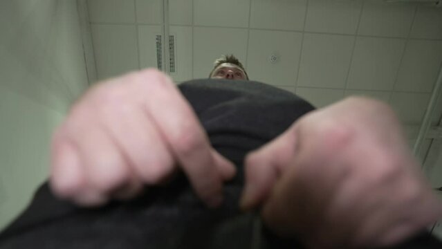 a young man with a beard and a hoodie and jeans appears in front of the camera and begins to unbuckle his belt and prepare to pee. shooting from the bottom point. A man in the toilet.