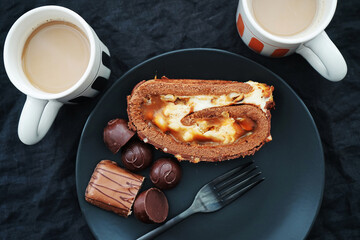 Chocolate roll with cream and caramel next to chocolate on a black plate near two cups of coffee with milk